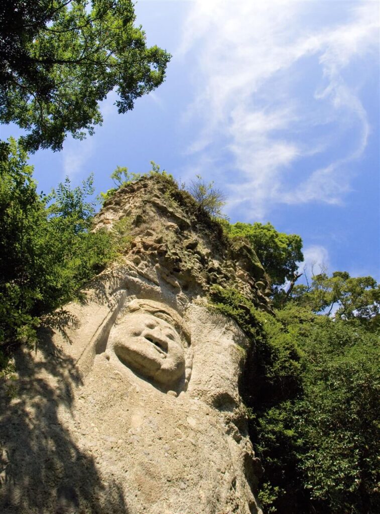 青空を背景に、森の中から見上げるようなアングルで撮影した大迫力の重要文化財・不動明王の磨崖仏