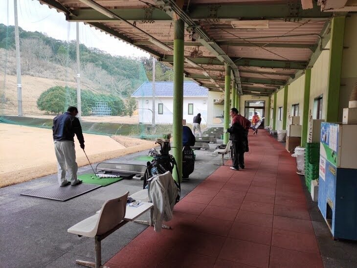 グリーンパーク直川ゴルフ練習場の打席エリア。屋根付きの広い打席に複数のゴルファーが並び、奥の白い建物と山を背景にショットを打っている