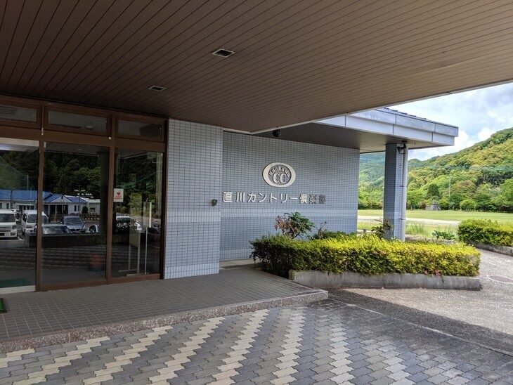 直川カントリー倶楽部クラブハウスの入り口。白いタイル壁に「直川カントリー倶楽部」のロゴが掲げられ、後方には山の緑が広がる